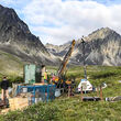 A drill tests for gold on a treeless area in front of rugged Alaska mountains.