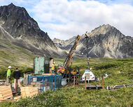 A drill tests for gold on a treeless area in front of rugged Alaska mountains.