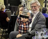 Young boy sitting on grandpa’s lap holds crystal award above his head.