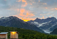 A sunset peeks over a mountain with the outhouses in the foreground.