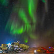 A mine crusher, conveyors, and trucks under a burst of green northern lights.
