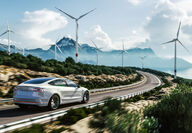 An EV drives past wind turbines on a rural road to the mountains.