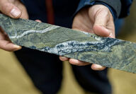 Hands holding a section of drill core with a quartz vein infilled with gold.