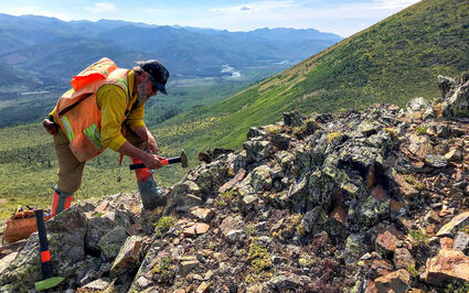 Geologist using a hammer to collect samples from a rock outcrop in Alaska.