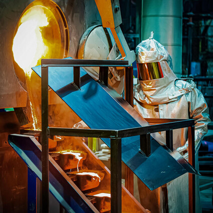 Gold poured from a kiln cascades down molds as worker in thermal suit looks on.