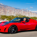 A red Tesla Roadster speeds past a large wind turbine farm.