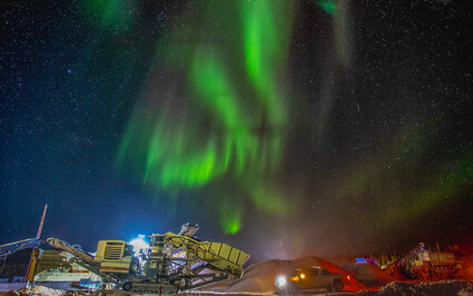 A mine crusher, conveyors, and trucks under a burst of green northern lights.